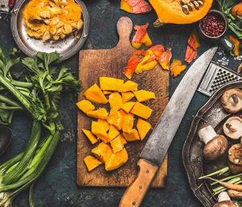 Diced pumpkin on a chopping board with a knife next to various vegetables