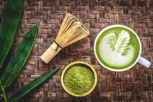 Image of Matcha Leaves next to bowl of matcha tea powder and a matcha latte