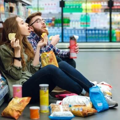 Man and woman sitting on supermarket floor eating crisps with crisp containers and bags on the floor