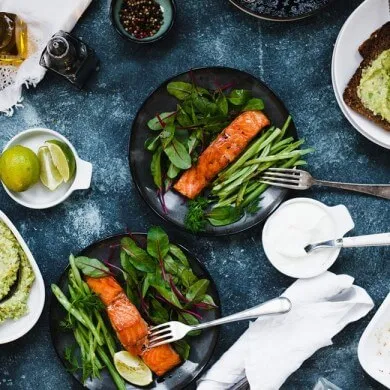 Two plates of grilled salmon with greens next to a tub of yogurt and bowl of slided lime